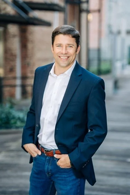 portrait of Jason Trew smiling; he is standing outside on a quiet urban street and wearing a sport coat, button down shirt, and jeans